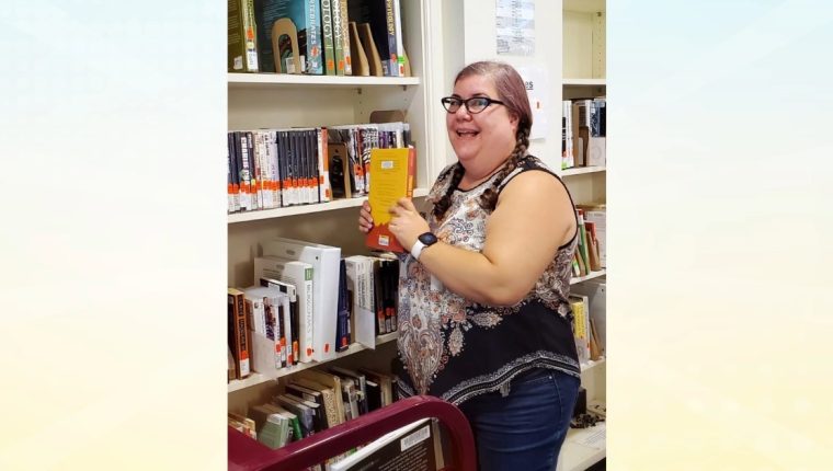A library worker stands in front of the course reserves with a textbook in hand